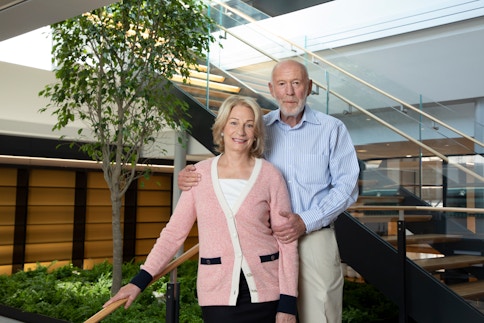 A photograph of Marilyn and Jim Simons. Marilyn is a woman with light blonde hair and is wearing a pink, black, and white button sweater with a white undershirt and black slacks. Jim Simons is a man with a faint white beard, a blue-and-white pinstripe button-down shirt, and beige slacks. They are situated in a room with a staircase leading upward and that has a small garden with a tree underneath it.